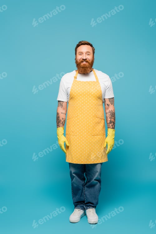 Preview: full length of bearded tattooed man in yellow apron and rubber gloves smiling at camera on blue