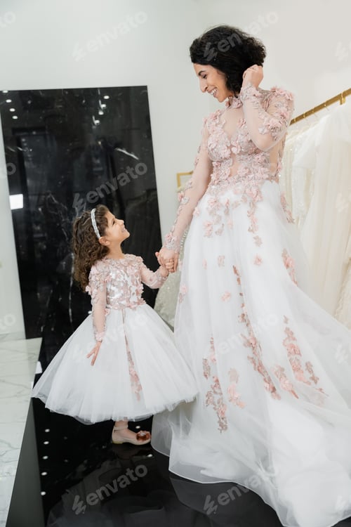 Preview: cheerful middle eastern woman with wavy hair in stunning wedding dress holding hands with daughter