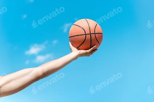 Preview: cropped shot of hand holding basketball ball against clear blue sky