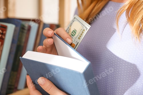 Preview: Woman hiding money in book indoors, closeup. Financial savings