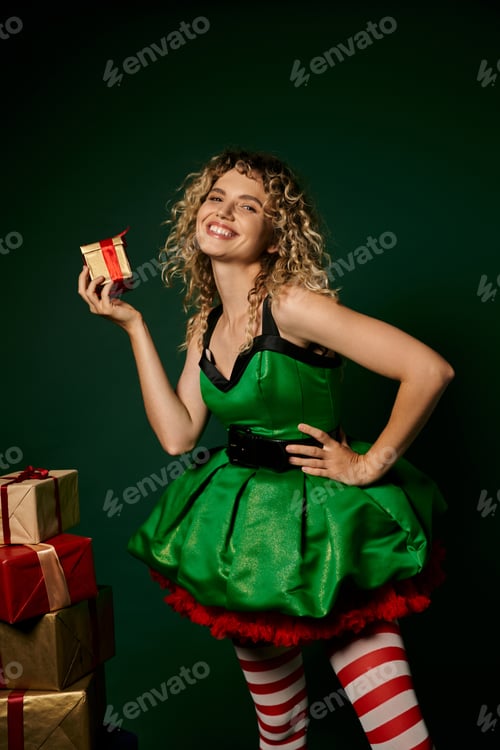Preview: jolly curly elf in festive dress posing with presents on dark green backdrop and looking at camera