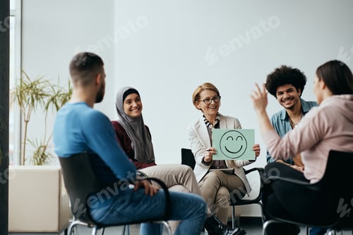 Preview: Happy mental health professional holds placard with drawn smiley face during group therapy meeting.