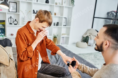Preview: overjoyed gay man covering mouth with hand near boyfriend making marriage proposal in bedroom