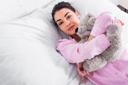 Preview: portrait of woman in pink pajamas with teddy bear resting in bed at home