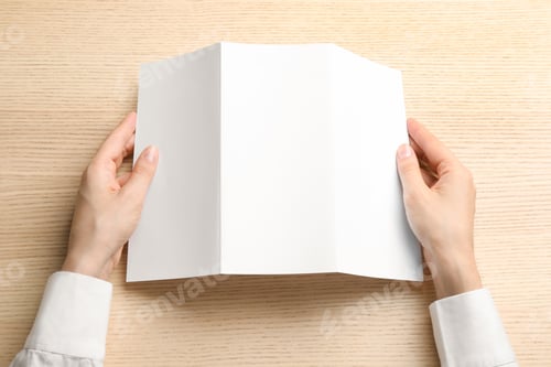 Preview: Young woman holding blank brochure at light wooden table, top view. Mock up for design
