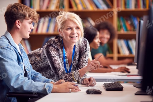Preview: Male University Or College Student Working At Computer In Library Being Helped By Tutor