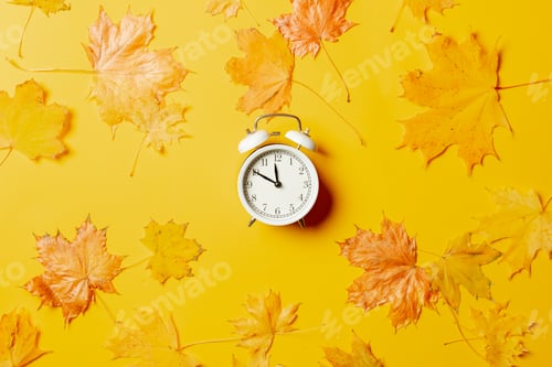 Preview: White alarm clock with maple leaves around on yellow background.