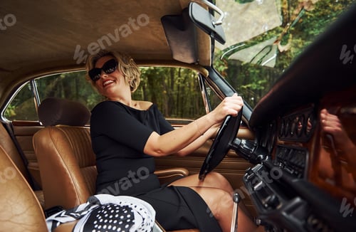 Preview: Blonde woman in sunglasses and in black dress sits in old vintage classic car