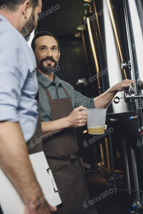 Preview: Male brewery worker pouring beer from the tank looking at his colleague