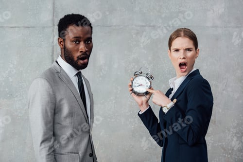 Preview: shocked businesswoman pointing at clock while african american businessman looking at camera on grey