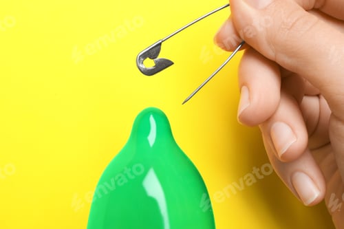 Preview: Woman piercing inflated condom with pin on yellow background, closeup. Safe sex