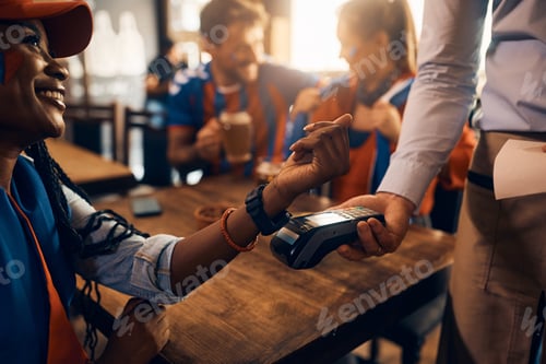 Preview: Close up of black woman paying contactless with smart watch during sports championship in bar.