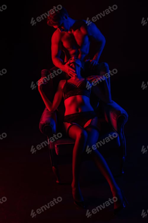 Preview: man touching sexy woman in underwear in armchair in red light isolated on black