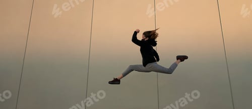 Preview: Athletic young woman with face mask dressed in black jumping in front of a gray wall.