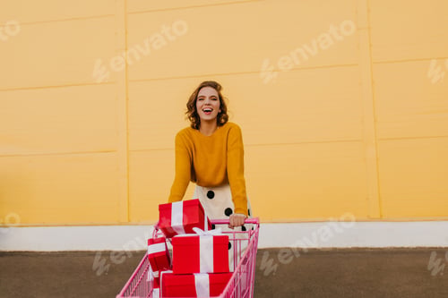 Preview: Satisfied girl posing with shopping cart full of presents. Outdoor photo of woman buying gifts