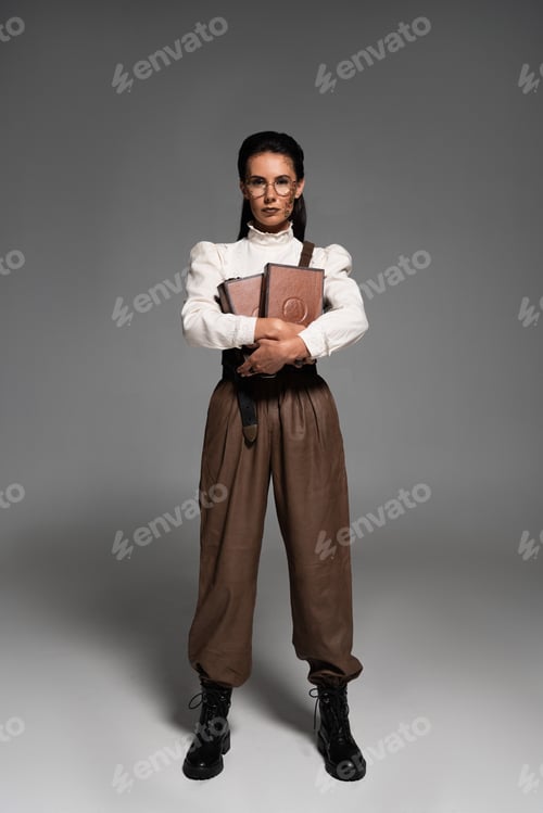 Preview: full length view of steampunk woman holding books and looking at camera on grey
