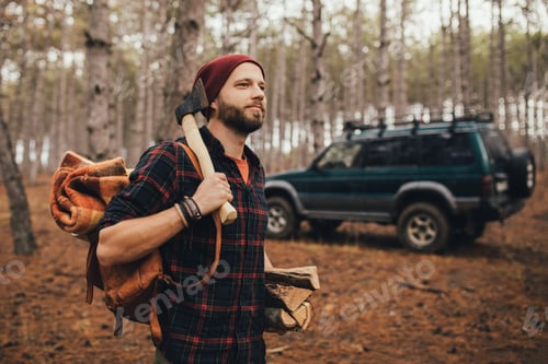 Preview: Millennial man hipster with backpack, holding axe and firewood in forest with car on background.