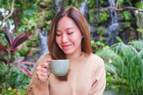 Preview: A young woman holding and drinking coffee while sitting in the garden with waterfall