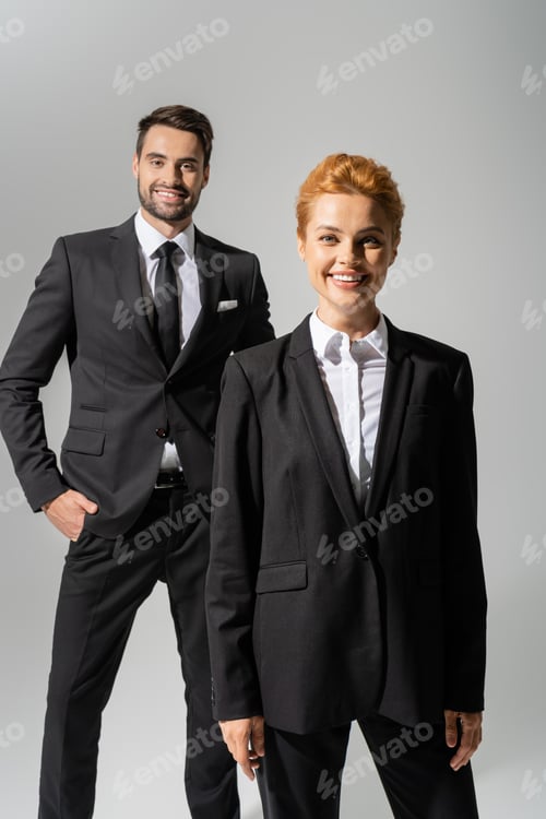 Preview: cheerful redhead businesswoman looking at camera near smiling colleague standing with hand in pocket