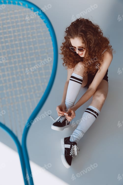 Preview: Smiling hipster girl in sunglasses sitting and tying shoelaces on grey