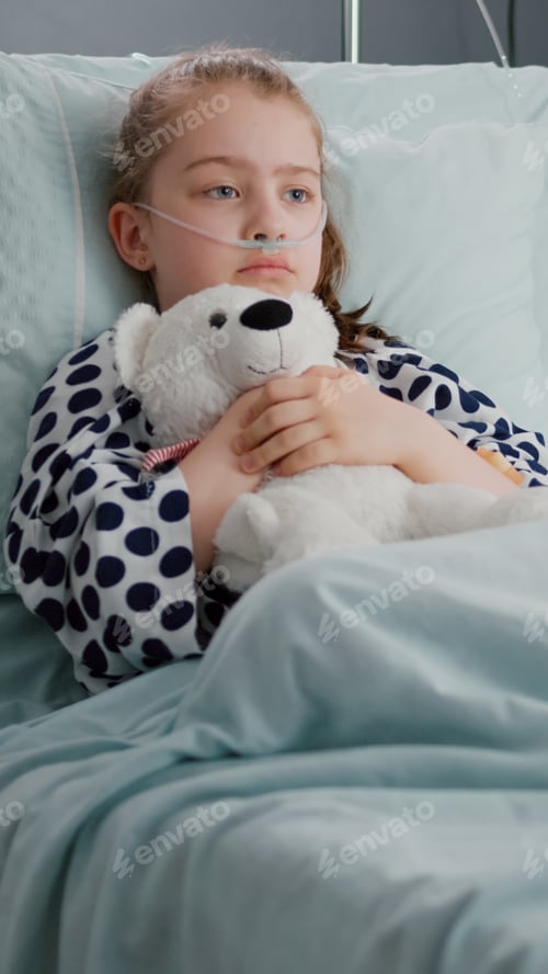 Preview: Alone worried child patient wearing oxygen nasal tube resting in bed holding teedy bear