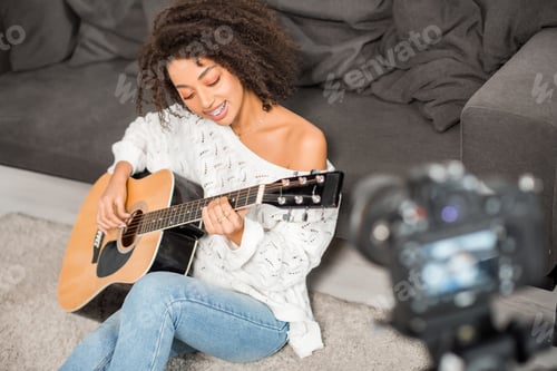 Preview: selective focus of happy african american girl in braces playing acoustic guitar near digital camera