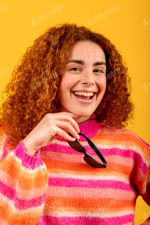 Preview: Redhead musician holding sunglasses and smiling on yellow background