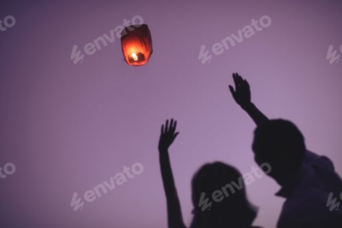 Preview: silhouettes of couple waving hands to flying sky lantern in evening
