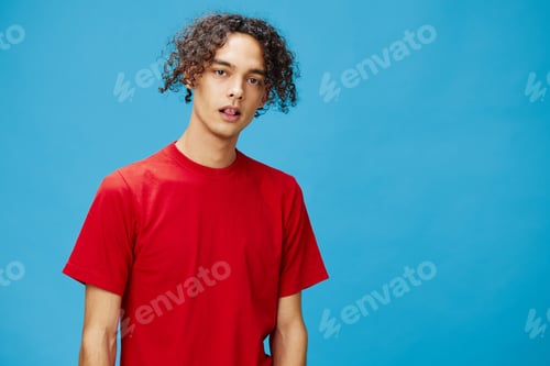 Preview: Attractive cute young curly tanned Caucasian guy in basic red t-shirt looks at camera posing