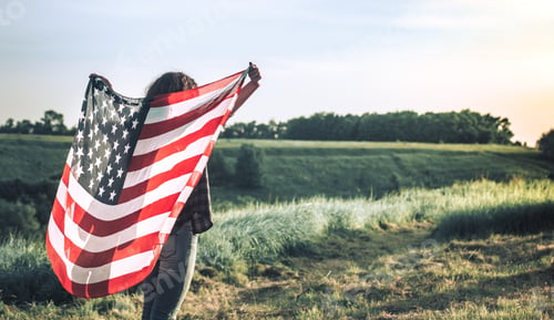 Preview: Young happy girl running and jumping carefree with open arms over wheat field. Holding USA flag.