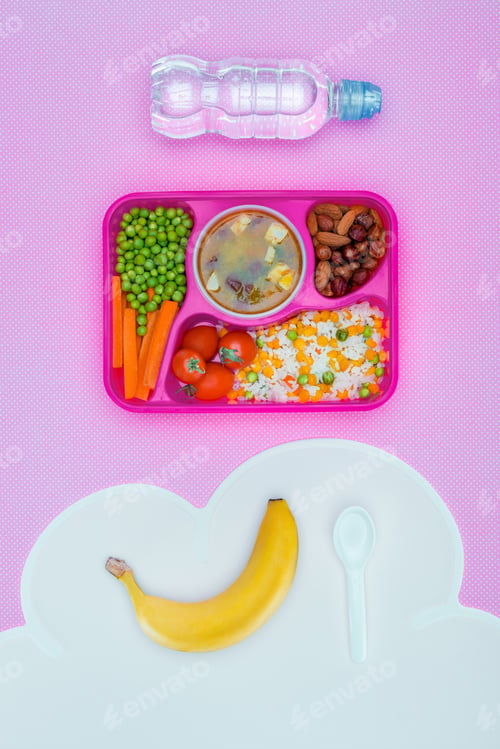 Preview: top view of tray with kids lunch for school, bottle of water and banana on violet table