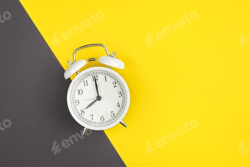 Preview: White alarm clock on a yellow and green background, flat lay.