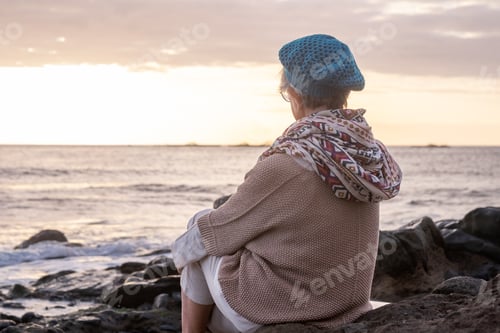 Preview: Rear view of relaxed caucasian senior woman sitting on the pebble beach at sunset light