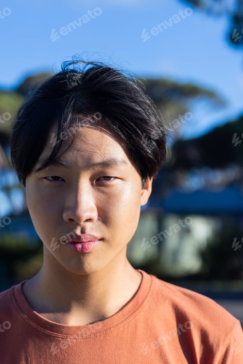 Preview: Asian male student hanging out outdoors, looking at camera with serious expression