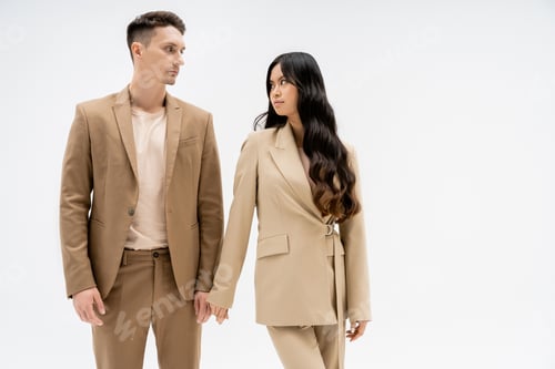Preview: fashionable multiethnic couple in beige suits looking at each other isolated on grey