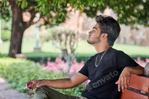 Preview: Attractive indian man smoker exhales cigarette smoke portrait in black t shirt and silver neck chain