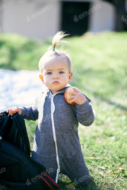 Preview: Little girl sits on her knees on a green lawn near a backpack and holds an apple in her hand