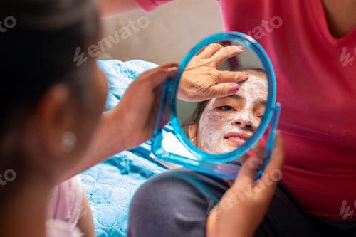 Preview: Latina woman looks in a mirror while her mother applies a face mask. Personal care incorporating.