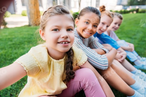 Preview: group of happy schoolgirls taking selfie while sitting on grass together