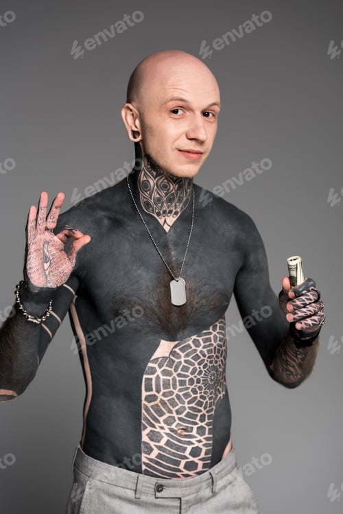 Preview: shirtless tattooed man with dollar banknotes showing ok sign and smiling at camera isolated on grey