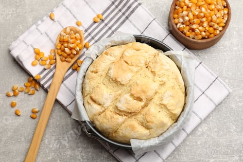 Preview: Freshly baked cornbread in cake pan, seeds and spoon on grey textured table, flat lay