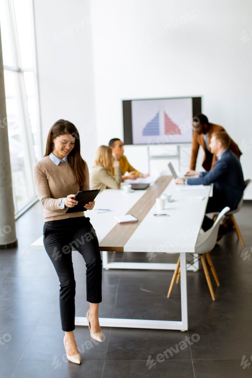 Preview: Young business woman standing in the office and using digital tablet in front of her team