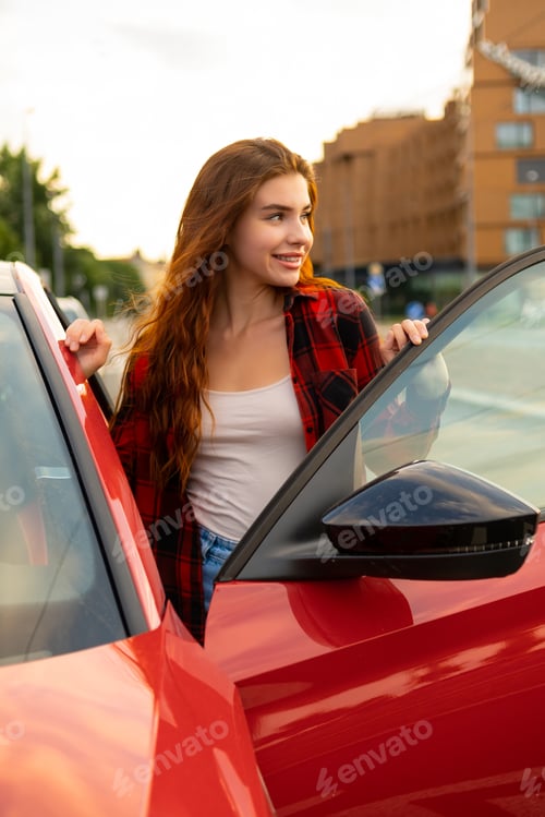 Preview: A smiling redhead woman in a plaid shirt stands next to a new red car