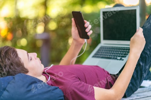 Preview: Teen boy with smartphone and laptop lies on the bench in the park.