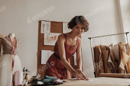 Preview: Curly young beautiful brunette woman in linen red dress with belt looks calm, works as fashion desi