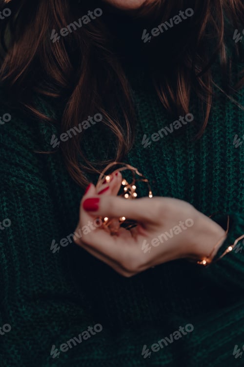 Preview: Young woman in green sweater and winter hat holds Christmas lights. Holiday time. Women’s hand