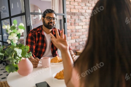 Preview: Happy couple eating breakfast and talking at dining table in morning. Indian girl and latino guy