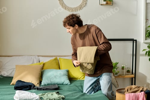 Preview: Handsome young man folding clothes in a cozy, modern bedroom during a sunny afternoon