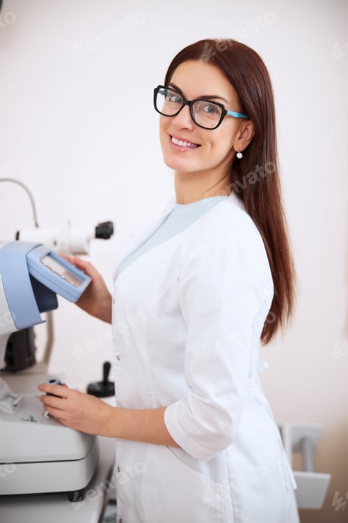 Preview: Beautiful eye doctor smiling while operating an auto refractometer