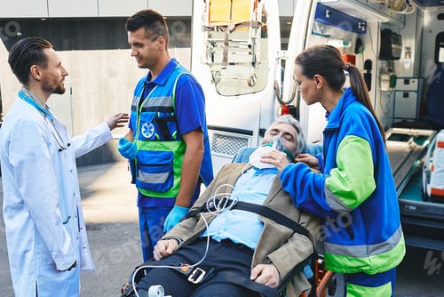 Preview: Doctor and paramedics checking health of mature male with breathing mask lying in ambulance gurney